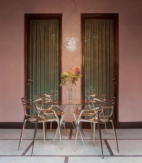 An artistic photo of a copper toned room with Kartell Masters Metal Chair in Copper shown around a transparent Dining Table